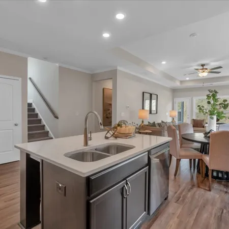 Modern kitchen with island sink, dining table, upholstered chairs, and open living space with stairs in background
