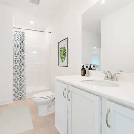 Bright modern bathroom with white vanity, shower with patterned curtain, toilet, and decorative plant artwork