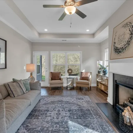 Bright living room with gray sofa, two armchairs, decorative rug, fireplace with art, and ceiling fan with light.