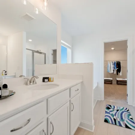 Bright modern bathroom with white vanity, large mirror, glass shower, and doorway to walk-in closet.