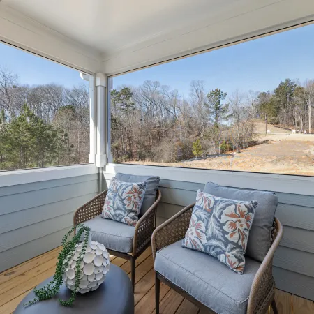 Cozy screened porch with two cushioned chairs, floral pillows, a small table, and a view of leafless trees.