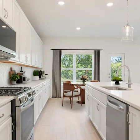 Modern white kitchen with large island, stainless steel appliances, wood chairs, and large windows with gray curtains.