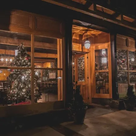 Cozy warmly lit restaurant exterior at night with decorated Christmas tree visible through large windows.