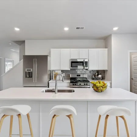Modern white kitchen with large island, three stools, stainless appliances, and bowl of fruit on countertop.