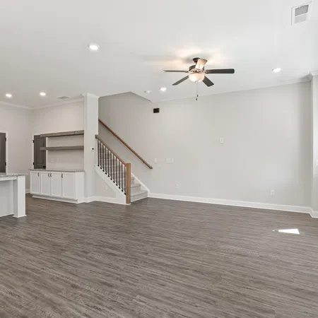 Spacious modern open living area with wood flooring, white walls, ceiling fan, stairs, and kitchen with stainless appliances.