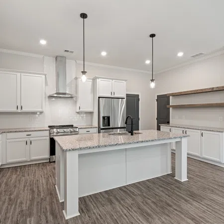 Modern white kitchen with large island, granite countertops, stainless steel appliances, and wood flooring under bright lights.
