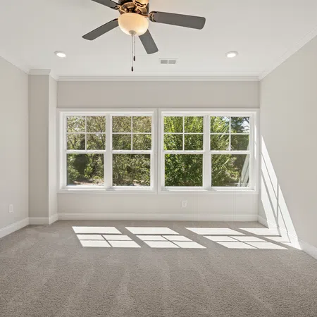 Empty bright room with beige carpet, large windows, white trim, and ceiling fan with light fixture.