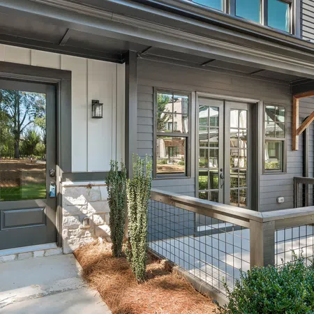 Modern townhouse entry with gray door, stone siding, wire railing porch, and landscaped greenery under bright daylight