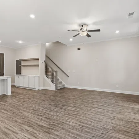 Spacious empty living room with wood flooring, ceiling fan, white kitchen island, and stairs in modern home