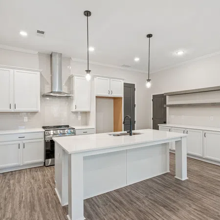 Modern white kitchen with large island, stainless steel stove, pendant lighting, and wood flooring in open layout.