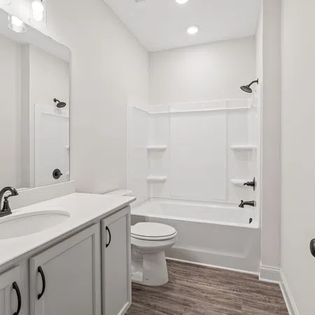 Modern white bathroom with bathtub, shower, toilet, gray cabinetry, and wood-look vinyl flooring under bright lighting