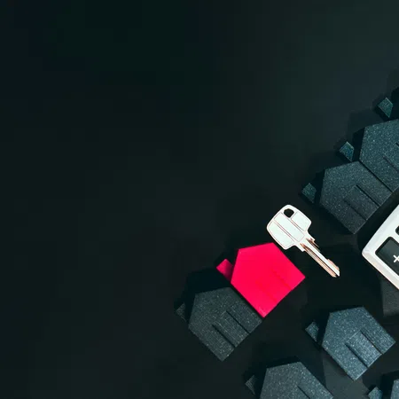 Calculator, house-shaped figures in black and red, and a silver key on a dark surface representing real estate finance.
