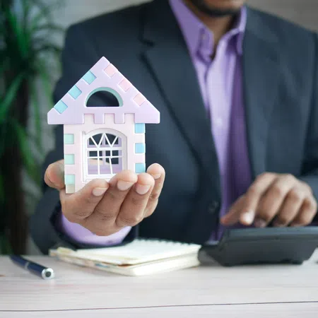 Businessman holding a small pastel-colored house model with notebook, pen, and calculator on desk.