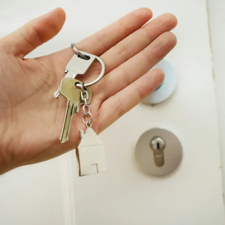 Hand holding house-shaped keychain and keys near a door lock symbolizing home ownership or real estate.