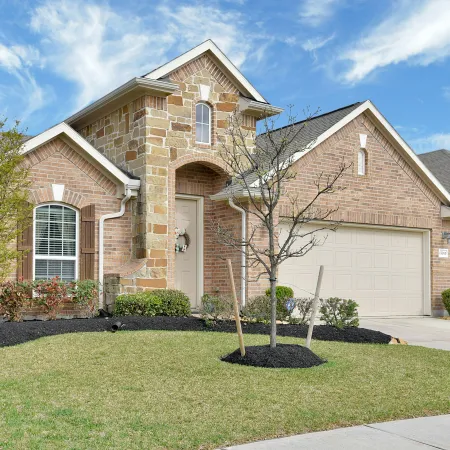 Brick and stone suburban house with a well-kept lawn, young trees, and blue sky with clouds overhead.
