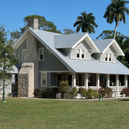 Gray craftsman-style house with stone chimney and large front porch surrounded by palm trees and greenery