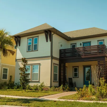 Two-story beige and cream house with wooden balcony and front porch in a sunny residential neighborhood