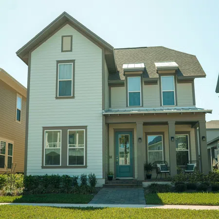 Two-story beige house with green door, front porch, and neatly trimmed lawn on a sunny day.