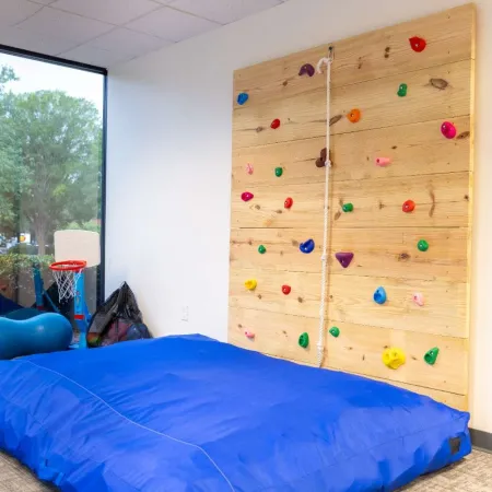 Indoor kids play area with a wooden climbing wall, blue crash mat, and basketball hoop near a large window.