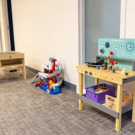 Children's play area with wooden toy workbench and colorful activity toys against a white wall in a carpeted room.