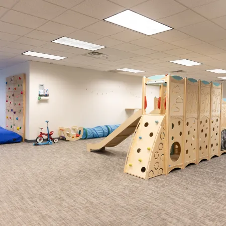 Bright indoor playroom with wooden climbing structure, slide, rock climbing wall, and toy vehicles on carpeted floor.