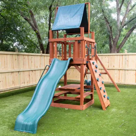 Wooden kids playset with blue slide, climbing wall, and canopy in fenced backyard with green grass.