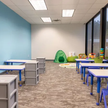 Bright classroom with blue and green chairs, tables, toys, and a play tent near large windows.