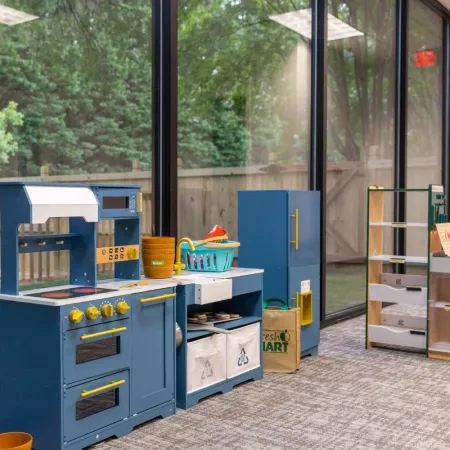 Children's play kitchen set with blue cabinets, toy food items, and play grocery shelves in a bright room.