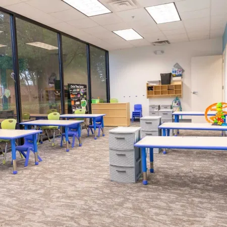 Bright classroom with blue tables, green chairs, toys, storage bins, and large windows overlooking trees.