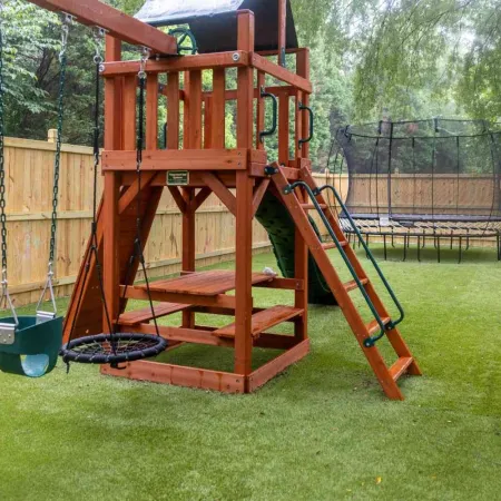 Children's wooden playground set with swings, slide, and trampoline on artificial grass in fenced backyard.