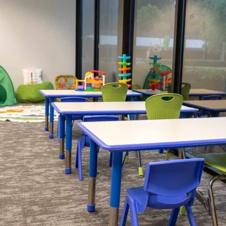 Bright children's classroom with blue and green chairs, white tables, and educational toys by large windows.