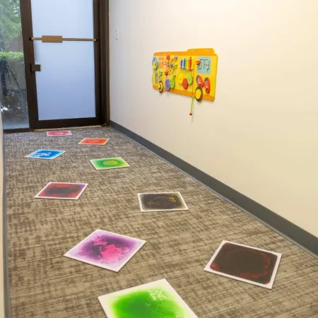 Colorful stepping stones arranged on a carpeted floor leading to a wall-mounted yellow activity board in a hallway.