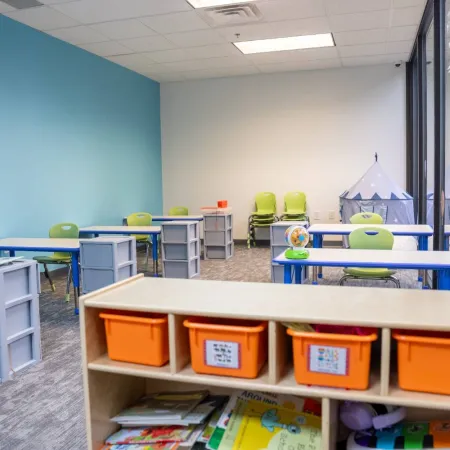 Bright classroom with blue and green desks, orange storage bins, and a windowed wall with educational posters.