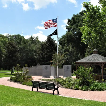 Peaceful park with American and POW flags, memorial stones, bench, gazebo, and green trees under blue sky.
