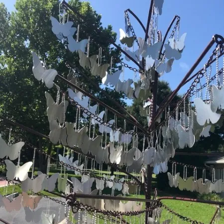 Metal butterfly shapes hanging from chains on a circular metal frame outdoors with trees and blue sky background