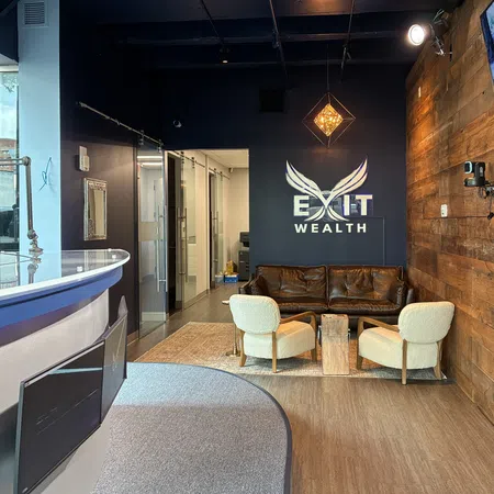 Modern office reception area with brown leather couch, white chairs, Exit Wealth logo, and wood paneled wall lighting.