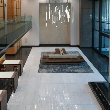 Modern office lobby with marble floors, central seating area on rug, pendant lighting, and glass balcony overlooking.