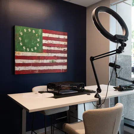 Modern office corner with white desk, ring light, American flag artwork with dollar signs, and large window view.