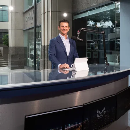 Man in blue suit standing behind modern news desk with microphone and tablet in studio with large windows.