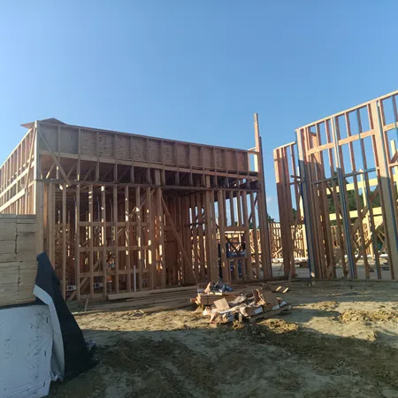 Wooden framing of a building under construction on a sunny day with stacks of lumber nearby