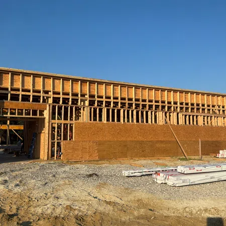Partially framed wooden building under construction on a sunny day with construction materials and gravel ground.