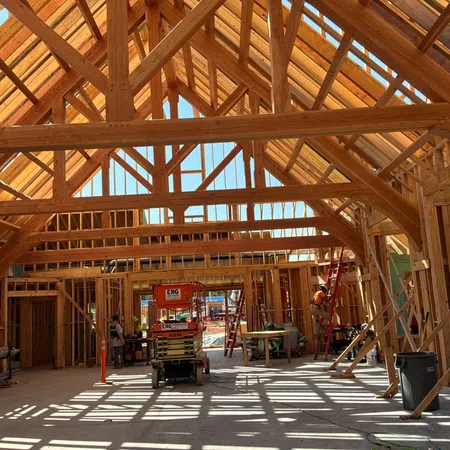 Interior view of a wooden building under construction with beams, rafters, and workers on site in daylight.