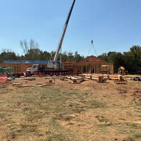 Crane lifting wooden roof frame at a construction site with building framework and scattered materials under clear sky.