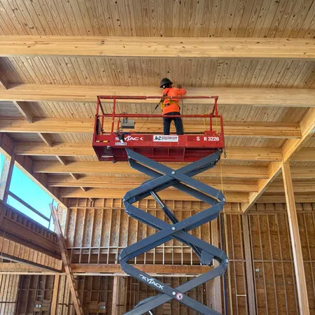 Construction worker in orange safety gear on elevated scissor lift working on wood ceiling beams inside building.