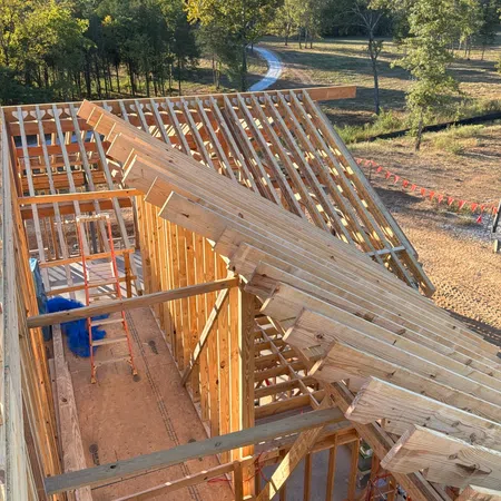 Wood frame construction of a house under bright daylight with roof trusses and surrounding trees in rural area.