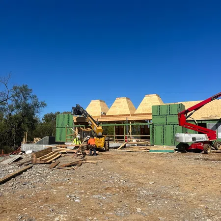 Construction site with workers, heavy machinery, and partially built wooden structures under a clear blue sky