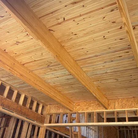 Interior view of wooden house framing with exposed beams and plywood ceiling under construction.