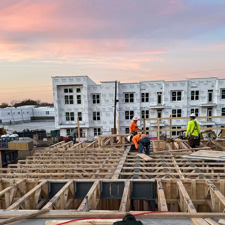 Construction workers building wooden framework of a residential development at sunset with white wrapped buildings in background.
