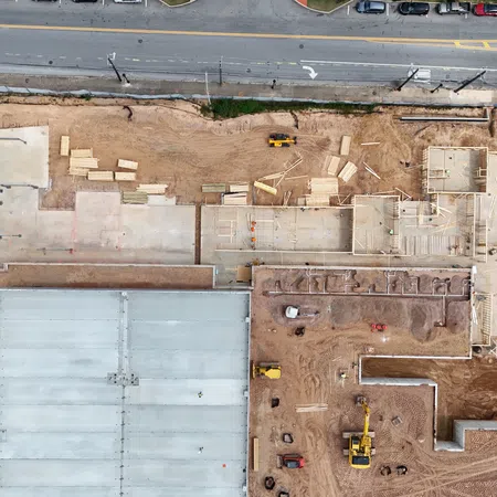 Aerial view of a construction site with foundation work, equipment, and surrounding streets and parked cars.