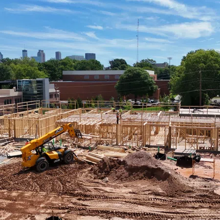 Construction site with wooden framing, heavy machinery, and workers building structures under a blue sky near urban buildings.
